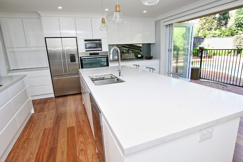 Huge trendy u-shaped light wood floor and brown floor kitchen pantry photo in Sydney with an undermount sink, flat-panel cabinets, white cabinets, quartz countertops, gray backsplash, glass sheet backsplash, stainless steel appliances and an island