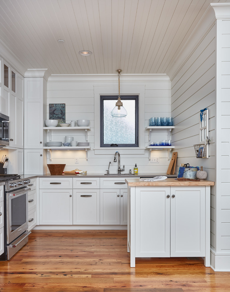 Exemple d'une cuisine bord de mer avec un placard à porte shaker, des portes de placard blanches, un électroménager en acier inoxydable, un sol en bois brun et fenêtre au-dessus de l'évier.