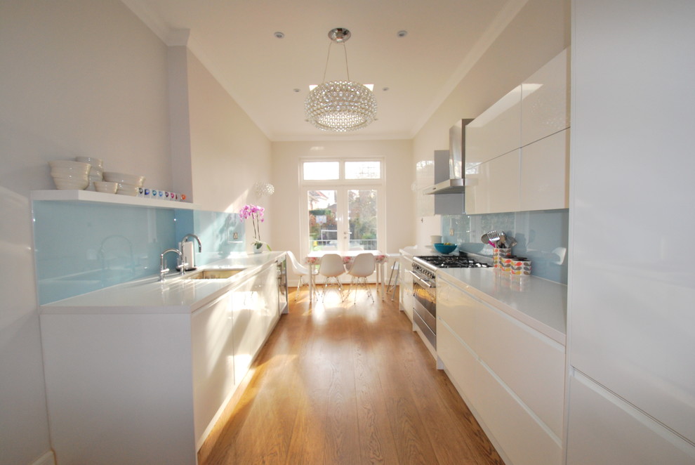Diseño de cocina comedor estrecha actual con armarios con paneles lisos, puertas de armario blancas, salpicadero azul, salpicadero de vidrio templado y encimera de acrílico