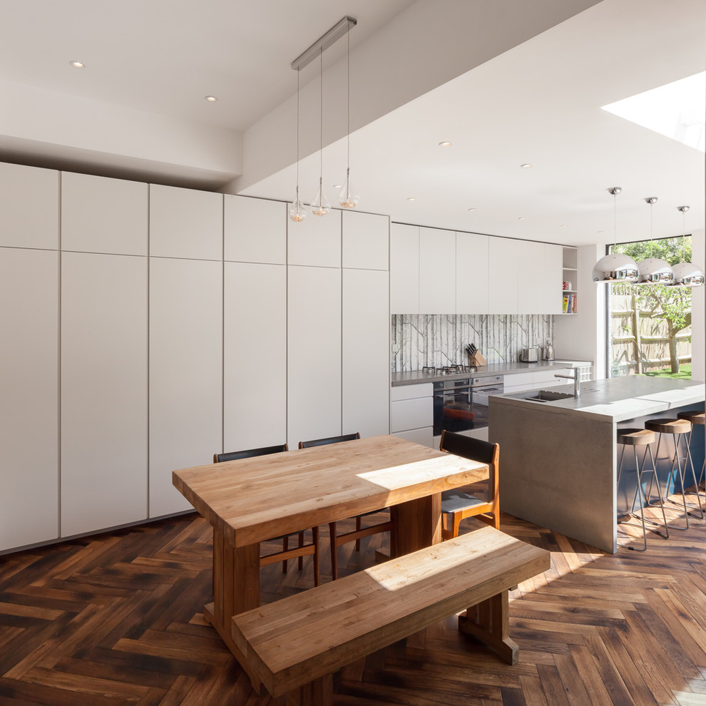 Cette image montre une cuisine américaine design avec un placard à porte plane, un plan de travail en béton, une crédence grise, un sol en bois brun, îlot et un sol marron.