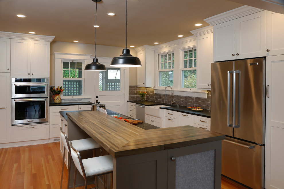 Design ideas for a classic kitchen/diner in Seattle with a belfast sink, shaker cabinets, white cabinets, composite countertops, brown splashback, metro tiled splashback, stainless steel appliances, light hardwood flooring and an island.