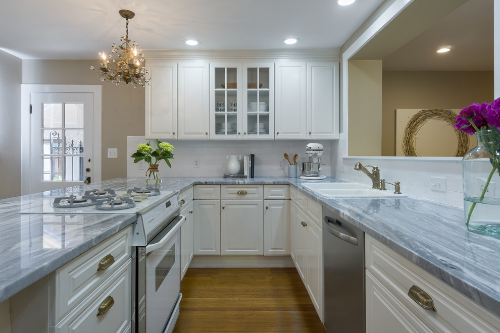 Small cottage chic u-shaped medium tone wood floor kitchen photo in Tampa with a drop-in sink, raised-panel cabinets, white cabinets, marble countertops, white backsplash, ceramic backsplash, white appliances and no island