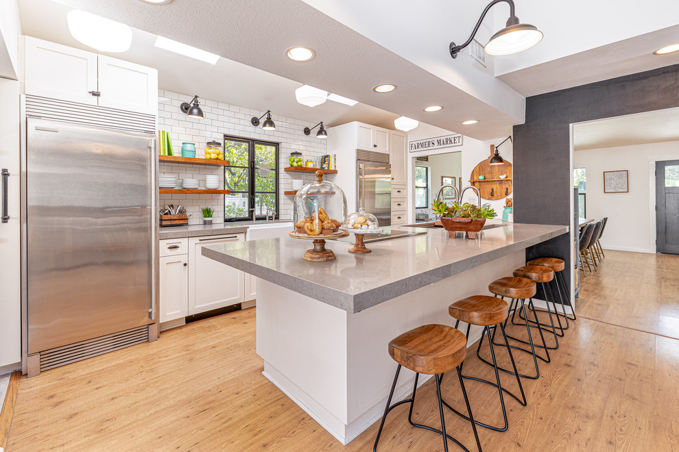 Country galley kitchen in Los Angeles with a belfast sink, shaker cabinets, white cabinets, white splashback, metro tiled splashback, stainless steel appliances, medium hardwood flooring, a breakfast bar, brown floors and grey worktops.