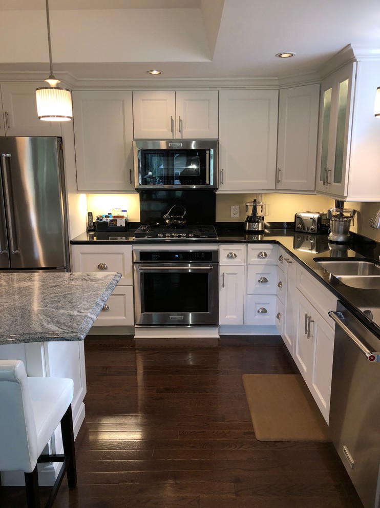 Large traditional l-shaped kitchen/diner in New York with a double-bowl sink, shaker cabinets, white cabinets, quartz worktops, beige splashback, stainless steel appliances, medium hardwood flooring, an island, brown floors and black worktops.