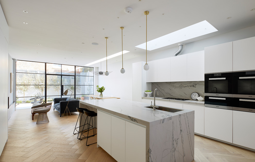 Medium sized contemporary single-wall open plan kitchen in Kent with a submerged sink, flat-panel cabinets, white cabinets, marble worktops, white splashback, marble splashback, integrated appliances, light hardwood flooring, a breakfast bar and beige floors.