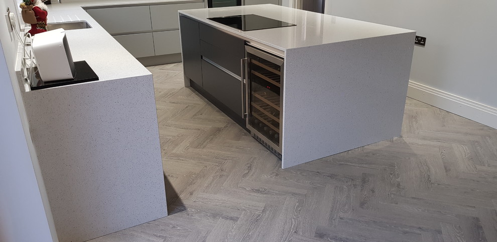 This is an example of a medium sized contemporary l-shaped kitchen/diner in Surrey with a submerged sink, flat-panel cabinets, grey cabinets, quartz worktops, white splashback, mirror splashback, stainless steel appliances, vinyl flooring, an island, brown floors and white worktops.