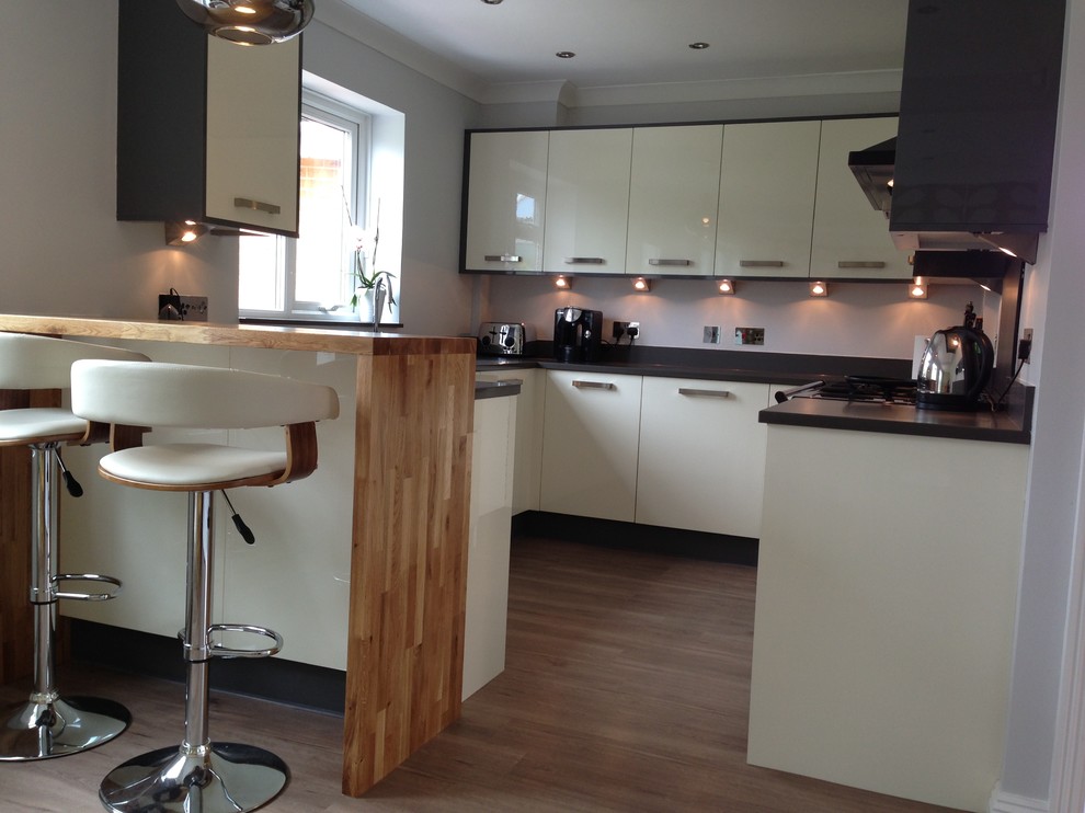 Photo of a medium sized contemporary grey and cream u-shaped kitchen/diner in Dorset with a submerged sink, flat-panel cabinets, beige cabinets, engineered stone countertops, metallic splashback, glass sheet splashback, stainless steel appliances, light hardwood flooring and a breakfast bar.