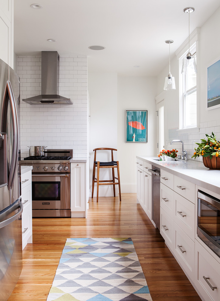 Modelo de cocina escandinava sin isla con fregadero bajoencimera, armarios estilo shaker, puertas de armario blancas, salpicadero blanco, salpicadero de azulejos tipo metro, electrodomésticos de acero inoxidable y suelo de madera en tonos medios