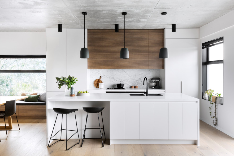 Bild på ett funkis vit vitt kök och matrum, med en dubbel diskho, släta luckor, vita skåp, grått stänkskydd, integrerade vitvaror, ljust trägolv, en köksö och beiget golv