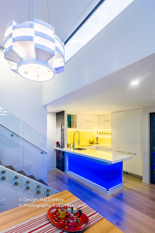 Inspiration for a mid-sized contemporary u-shaped light wood floor eat-in kitchen remodel in Auckland with an undermount sink, flat-panel cabinets, white cabinets, solid surface countertops, yellow backsplash, glass sheet backsplash, white appliances and no island