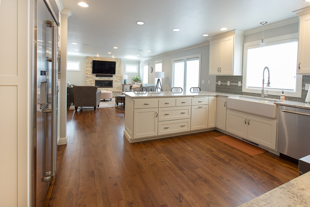 Example of a mid-sized transitional l-shaped medium tone wood floor eat-in kitchen design in Other with a farmhouse sink, shaker cabinets, white cabinets, granite countertops, gray backsplash, subway tile backsplash, stainless steel appliances and a peninsula