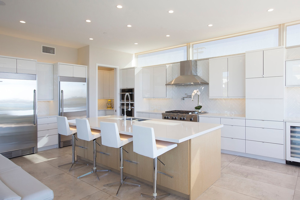 Photo of a contemporary l-shaped open plan kitchen in San Diego with a double-bowl sink, flat-panel cabinets, white cabinets, white splashback, metro tiled splashback, stainless steel appliances, an island and beige floors.