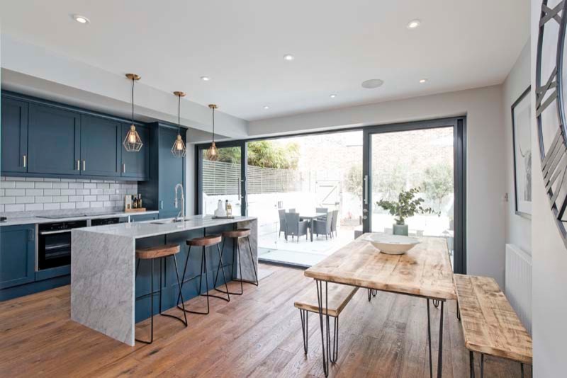 Inspiration for a medium sized contemporary galley open plan kitchen in London with a built-in sink, shaker cabinets, blue cabinets, marble worktops, white splashback, metro tiled splashback, black appliances, dark hardwood flooring and an island.