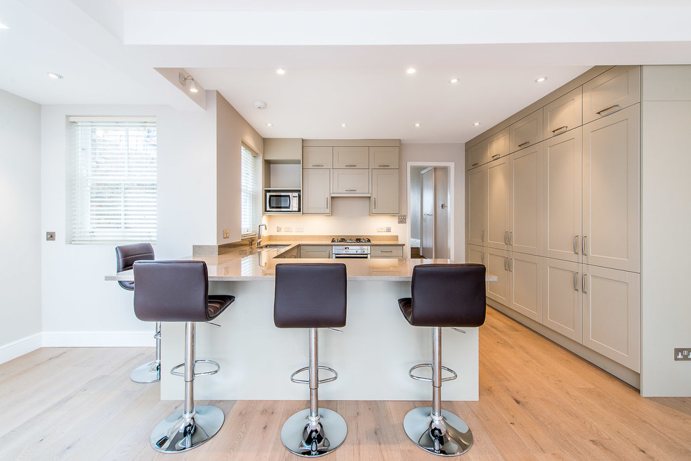 This is an example of a traditional kitchen in London with a submerged sink, shaker cabinets, grey cabinets, stainless steel appliances, light hardwood flooring and a breakfast bar.