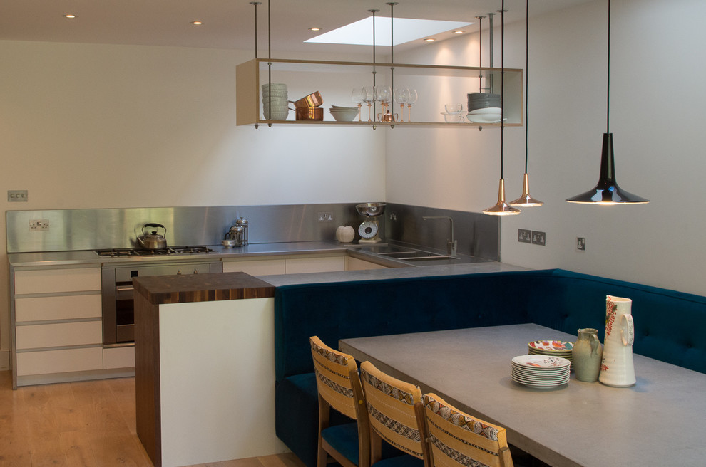 Medium sized contemporary u-shaped open plan kitchen in London with a built-in sink, flat-panel cabinets, white cabinets, wood worktops, metallic splashback, black appliances, light hardwood flooring, no island and brown worktops.