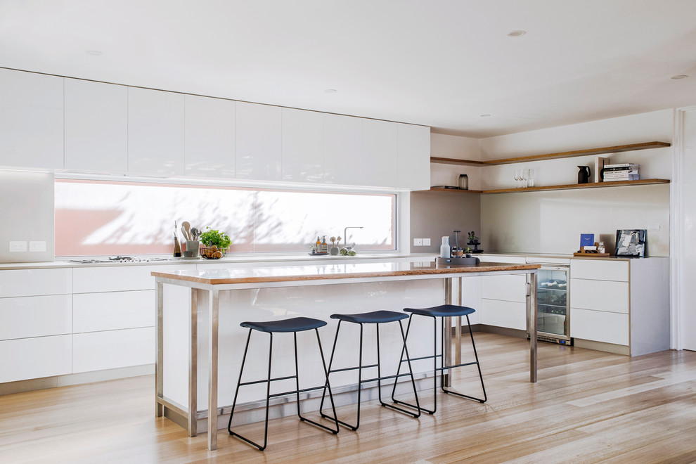 Exempel på ett modernt kök, med släta luckor, vita skåp, fönster som stänkskydd, rostfria vitvaror, ljust trägolv, en köksö och brunt golv