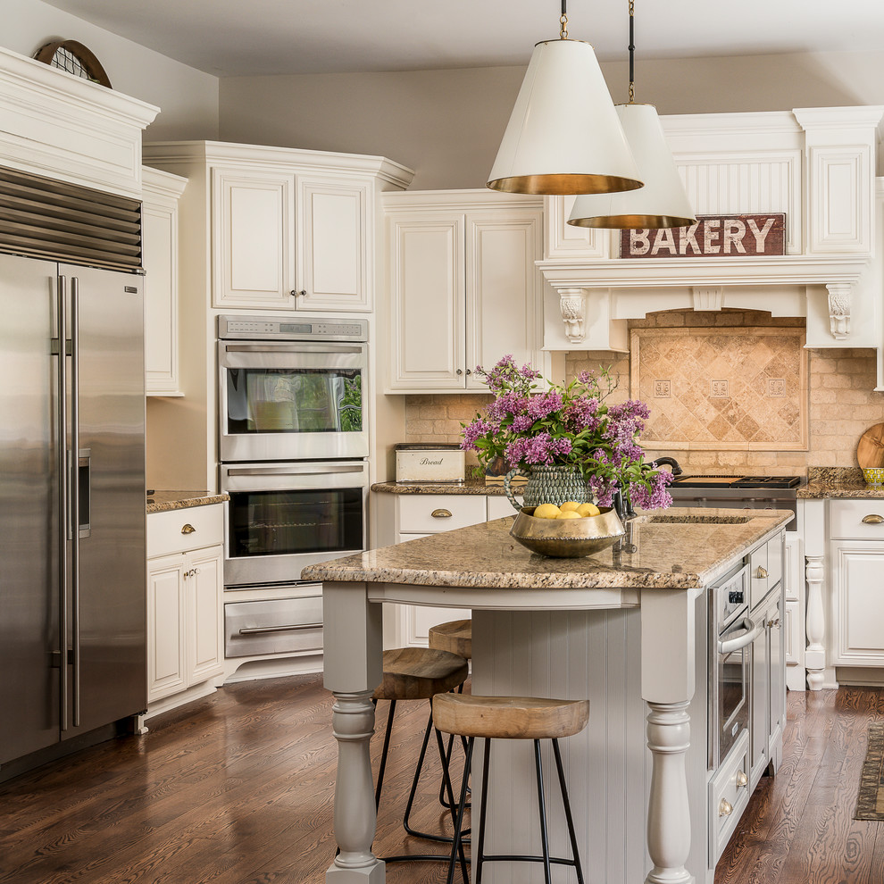 Klassisk inredning av ett kök, med luckor med upphöjd panel, vita skåp, beige stänkskydd, rostfria vitvaror, mellanmörkt trägolv, en köksö och brunt golv
