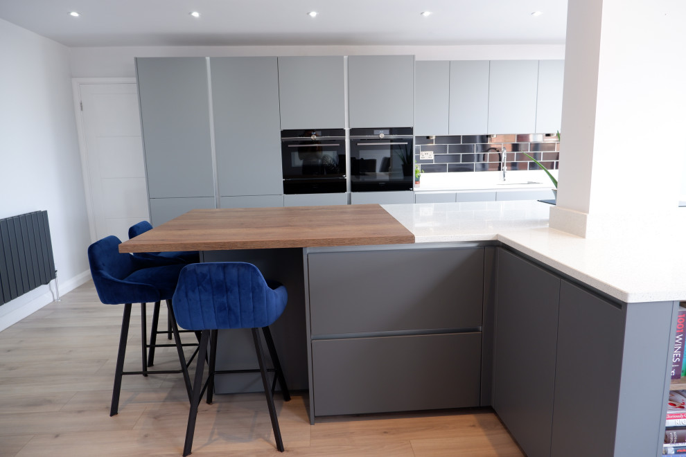 Design ideas for a large modern single-wall kitchen/diner in Other with an integrated sink, flat-panel cabinets, grey cabinets, quartz worktops, black splashback, metro tiled splashback, black appliances, laminate floors, an island, brown floors and white worktops.