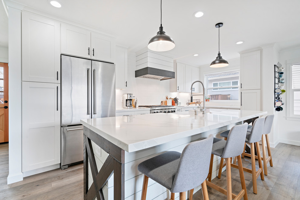 Farmhouse l-shaped medium tone wood floor and brown floor kitchen photo in San Francisco with a farmhouse sink, shaker cabinets, white cabinets, white backsplash, stainless steel appliances, an island and white countertops