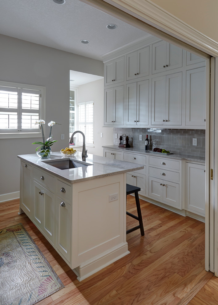 Photo of a small beach style galley enclosed kitchen in Jacksonville with a single-bowl sink, beaded cabinets, white cabinets, engineered stone countertops, grey splashback, porcelain splashback, stainless steel appliances, light hardwood flooring and an island.