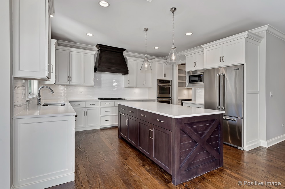 Example of a large arts and crafts u-shaped light wood floor enclosed kitchen design in Chicago with an undermount sink, shaker cabinets, white cabinets, solid surface countertops, white backsplash, subway tile backsplash, stainless steel appliances and an island