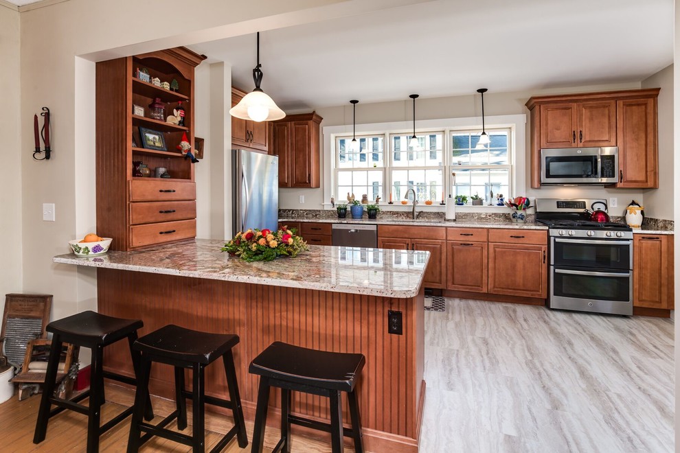 Photo of a medium sized country u-shaped kitchen/diner in Providence with a submerged sink, raised-panel cabinets, brown cabinets, granite worktops, beige splashback, stainless steel appliances, vinyl flooring, a breakfast bar and grey floors.