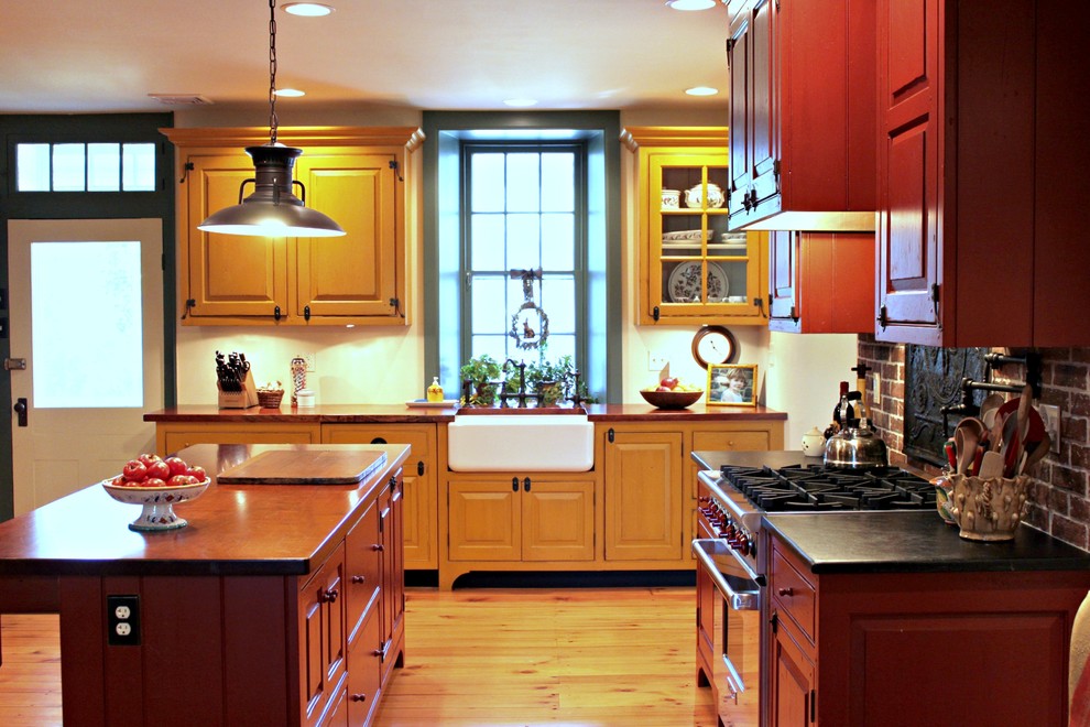 Inredning av ett lantligt mellanstort kök, med en rustik diskho, luckor med upphöjd panel, skåp i mellenmörkt trä, bänkskiva i täljsten, rött stänkskydd, rostfria vitvaror, ljust trägolv och en köksö