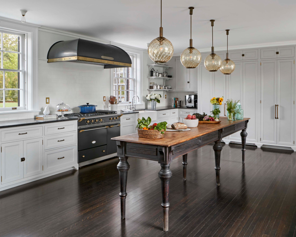 Elegant l-shaped medium tone wood floor and brown floor enclosed kitchen photo in Other with a farmhouse sink, recessed-panel cabinets, white cabinets, white backsplash, ceramic backsplash, black appliances, an island and black countertops
