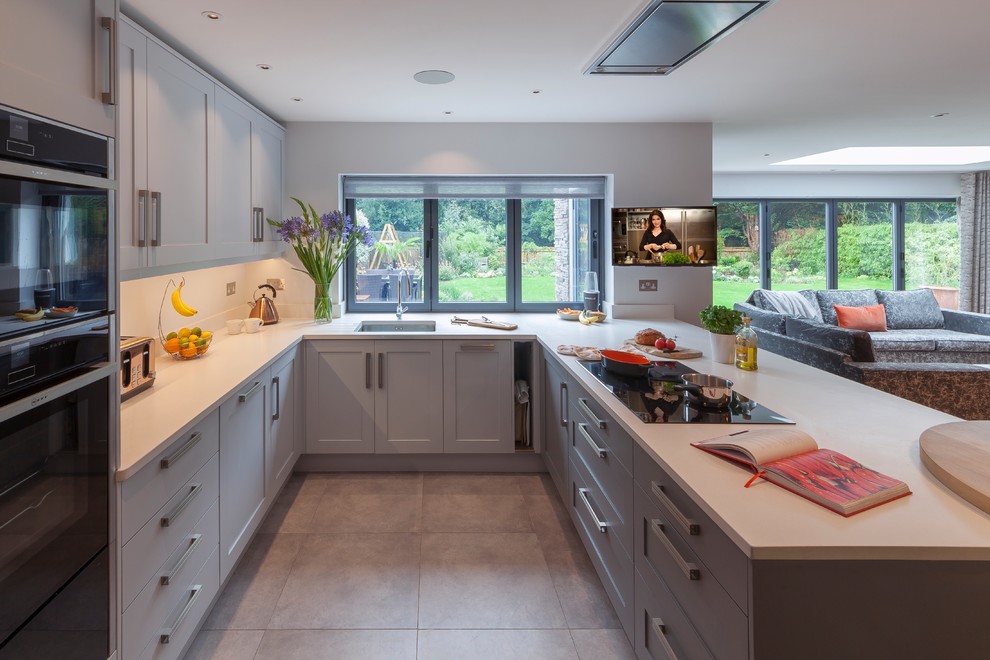 Medium sized traditional u-shaped open plan kitchen in Surrey with shaker cabinets, grey cabinets, composite countertops, white splashback, porcelain flooring, a breakfast bar, grey floors, a submerged sink and black appliances.