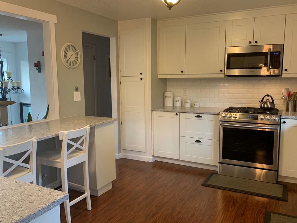 Bild på ett mellanstort vintage grå grått kök, med en dubbel diskho, skåp i shakerstil, vita skåp, bänkskiva i kvartsit, vitt stänkskydd, stänkskydd i tunnelbanekakel, rostfria vitvaror, laminatgolv, en halv köksö och brunt golv