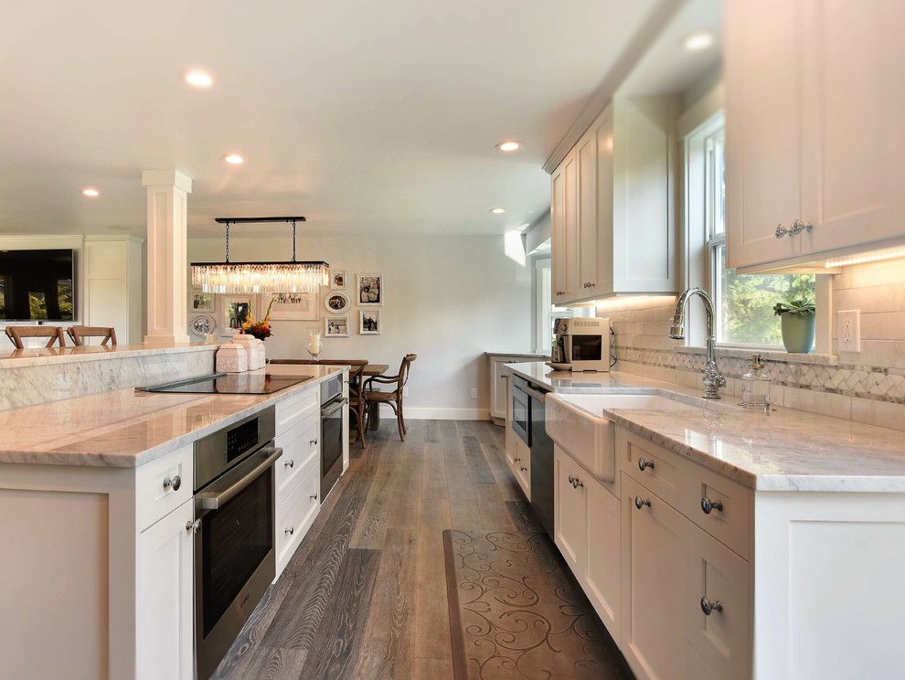 Eat-in kitchen - mid-sized transitional galley medium tone wood floor eat-in kitchen idea in Seattle with a farmhouse sink, shaker cabinets, white cabinets, marble countertops, white backsplash, stone tile backsplash, stainless steel appliances and an island