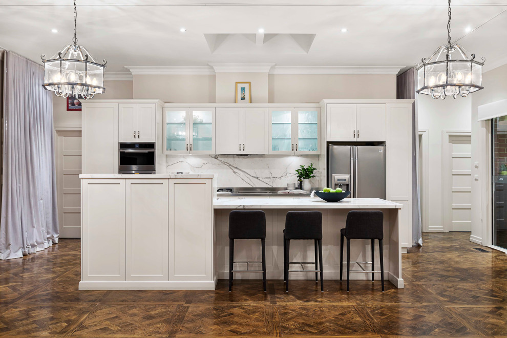 This is an example of a large traditional galley open plan kitchen in Melbourne with shaker cabinets, white cabinets, marble worktops, white splashback, marble splashback, stainless steel appliances, an island, brown floors, white worktops and dark hardwood flooring.