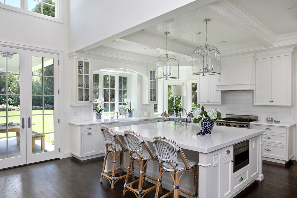 Elegant l-shaped dark wood floor and brown floor kitchen photo in San Francisco with a farmhouse sink, shaker cabinets, white cabinets, white backsplash, subway tile backsplash, stainless steel appliances, an island and white countertops