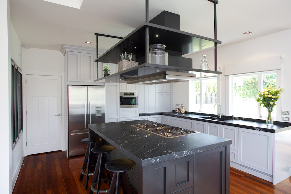 Example of a mid-sized classic l-shaped medium tone wood floor and brown floor eat-in kitchen design in Auckland with a double-bowl sink, recessed-panel cabinets, gray cabinets, quartzite countertops, black backsplash, stone slab backsplash, stainless steel appliances and an island