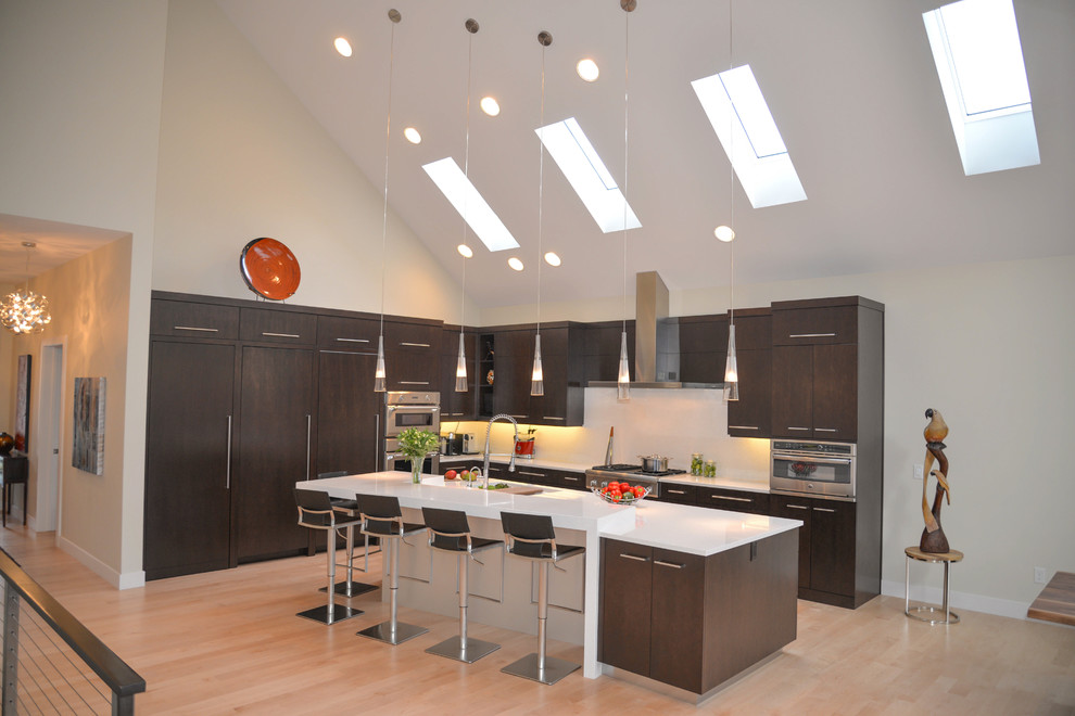 Example of a large trendy l-shaped light wood floor kitchen pantry design in Grand Rapids with a double-bowl sink, flat-panel cabinets, dark wood cabinets, solid surface countertops, white backsplash, stone slab backsplash, stainless steel appliances and an island