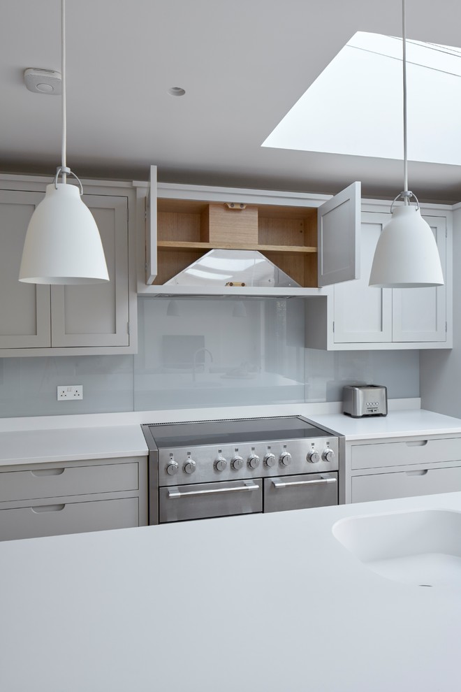 This is an example of a medium sized contemporary single-wall open plan kitchen in Surrey with an integrated sink, shaker cabinets, grey cabinets, engineered stone countertops, white splashback, glass sheet splashback, stainless steel appliances, light hardwood flooring, an island and beige floors.