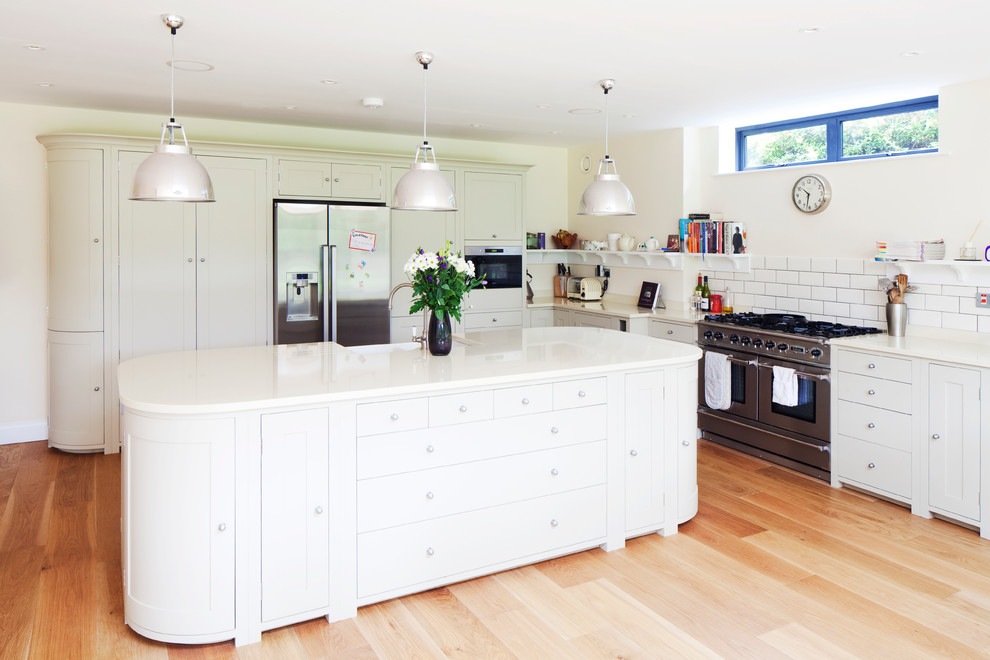 Klassische Küche in L-Form mit Landhausspüle, Schrankfronten im Shaker-Stil, weißen Schränken, Küchenrückwand in Weiß, Rückwand aus Metrofliesen, Küchengeräten aus Edelstahl, braunem Holzboden und Kücheninsel in Sussex