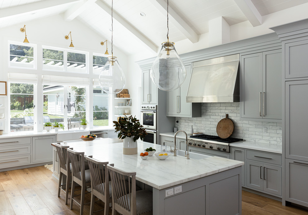 Inspiration pour une grande cuisine traditionnelle en L avec plan de travail en marbre, un électroménager en acier inoxydable, un sol en bois brun, îlot, un évier de ferme, un placard à porte shaker, des portes de placard grises, une crédence blanche, une crédence en marbre, un plan de travail blanc et fenêtre au-dessus de l'évier.