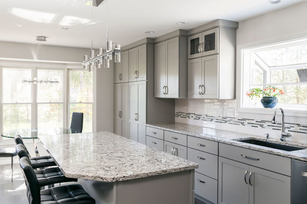 Large contemporary l-shaped kitchen pantry in St Louis with a single-bowl sink, recessed-panel cabinets, grey cabinets, engineered stone countertops, white splashback, glass tiled splashback, stainless steel appliances, vinyl flooring, an island, grey floors and grey worktops.