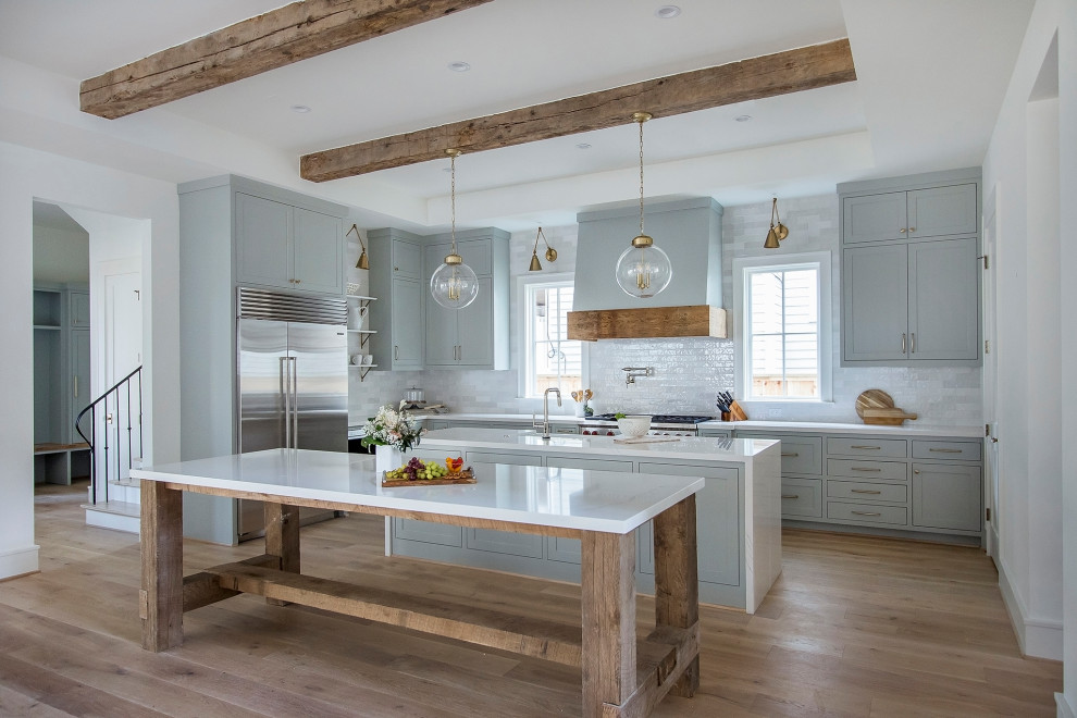 Inspiration for a country l-shaped kitchen/diner in Houston with a belfast sink, shaker cabinets, grey cabinets, white splashback, metro tiled splashback, medium hardwood flooring, an island, brown floors and white worktops.