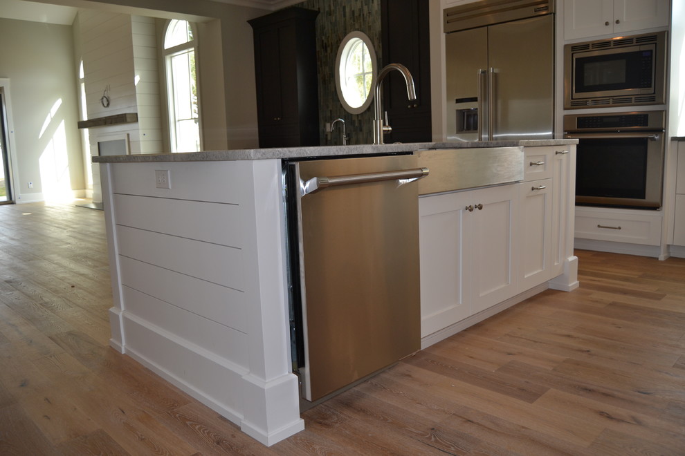 Example of a mid-sized beach style medium tone wood floor eat-in kitchen design in Jacksonville with a farmhouse sink, recessed-panel cabinets, white cabinets, quartz countertops, white backsplash, glass tile backsplash, stainless steel appliances and an island