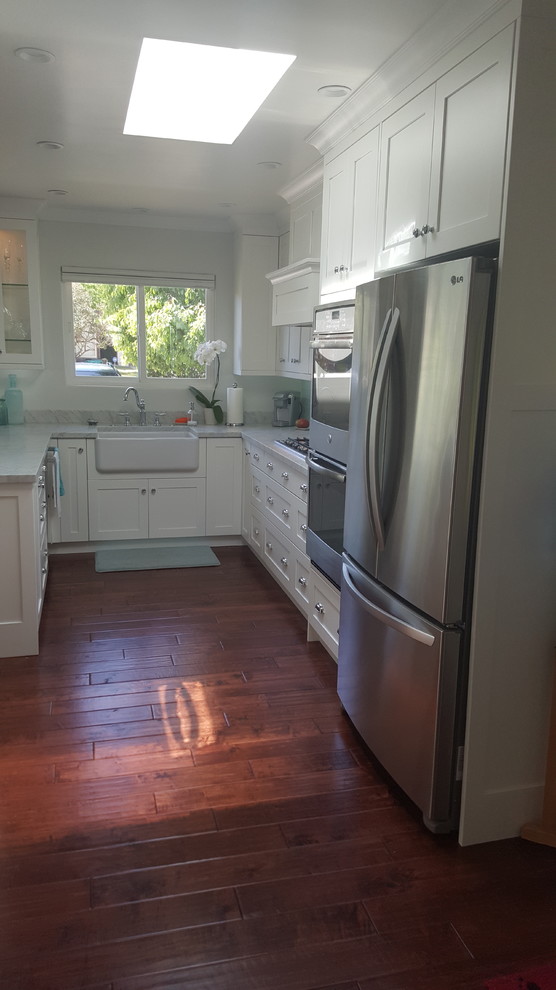 This is an example of a medium sized nautical u-shaped kitchen/diner in Los Angeles with a belfast sink, shaker cabinets, white cabinets, marble worktops, blue splashback, glass tiled splashback, stainless steel appliances, dark hardwood flooring, a breakfast bar and brown floors.