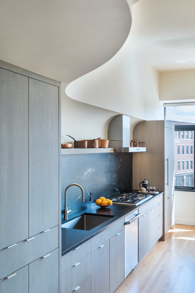 Example of a mid-sized eclectic single-wall light wood floor open concept kitchen design in Boston with an undermount sink, flat-panel cabinets, light wood cabinets, soapstone countertops, black backsplash, stone slab backsplash, paneled appliances and an island