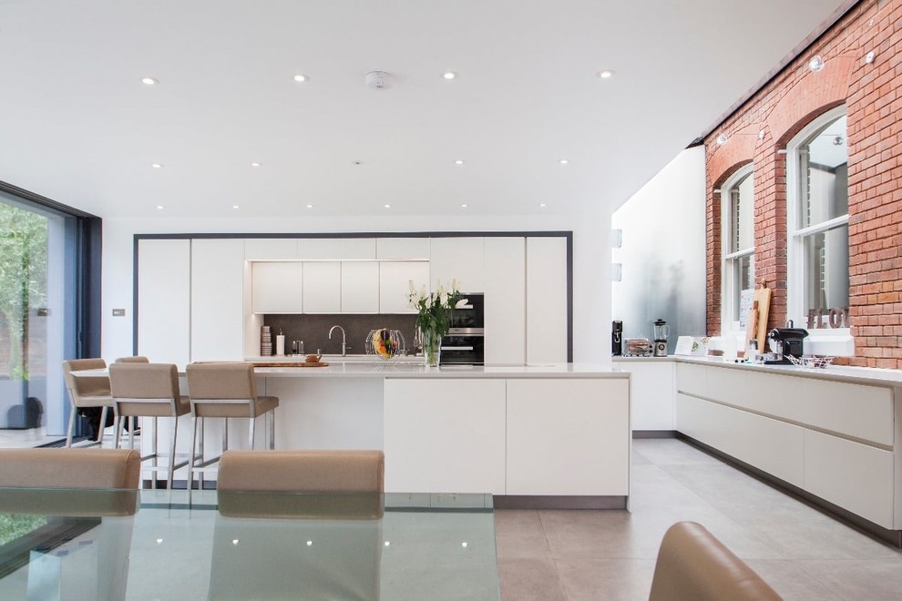 Photo of a large contemporary l-shaped kitchen in Hampshire with a triple-bowl sink, flat-panel cabinets, white cabinets, grey splashback, stone tiled splashback, black appliances, ceramic flooring, an island and grey floors.