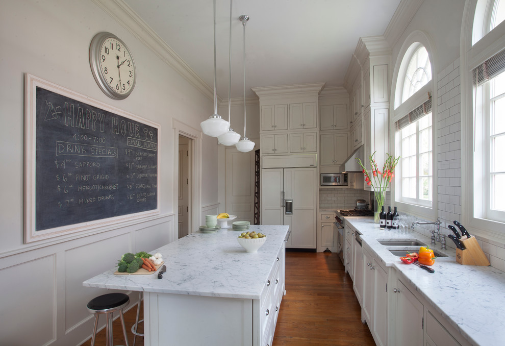 Example of a classic l-shaped medium tone wood floor enclosed kitchen design in New Orleans with a double-bowl sink, shaker cabinets, white cabinets, white backsplash, subway tile backsplash, paneled appliances and an island