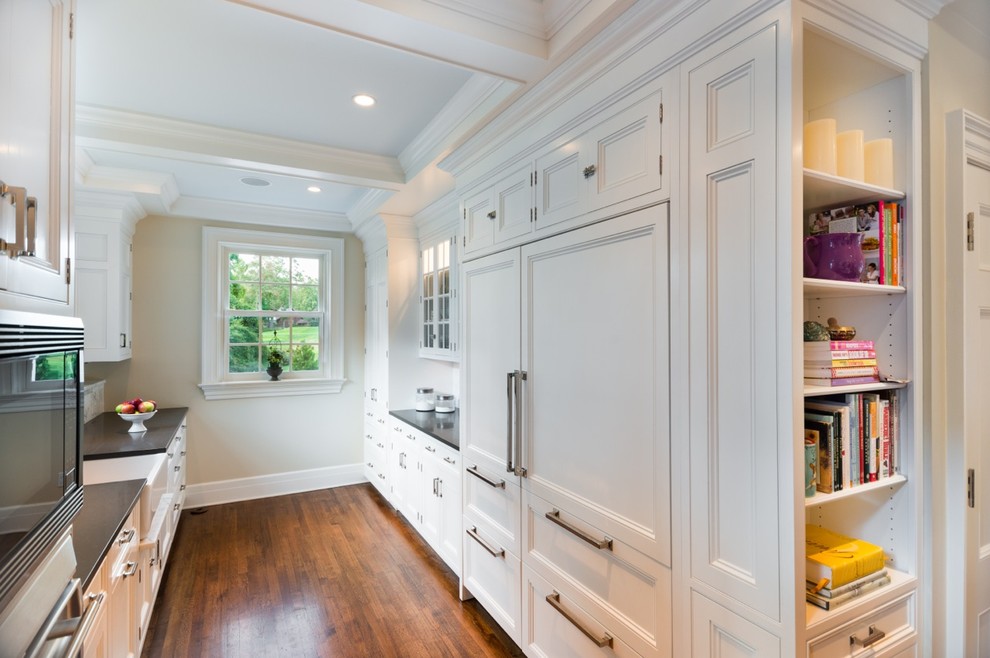 This is an example of a traditional galley kitchen/diner in New York with a belfast sink, recessed-panel cabinets, white cabinets, engineered stone countertops, white splashback, stone tiled splashback and integrated appliances.