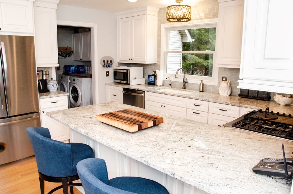 Klassisk inredning av ett grå grått kök, med en undermonterad diskho, vita skåp, granitbänkskiva, flerfärgad stänkskydd, stänkskydd i porslinskakel, rostfria vitvaror, ljust trägolv, en halv köksö och brunt golv