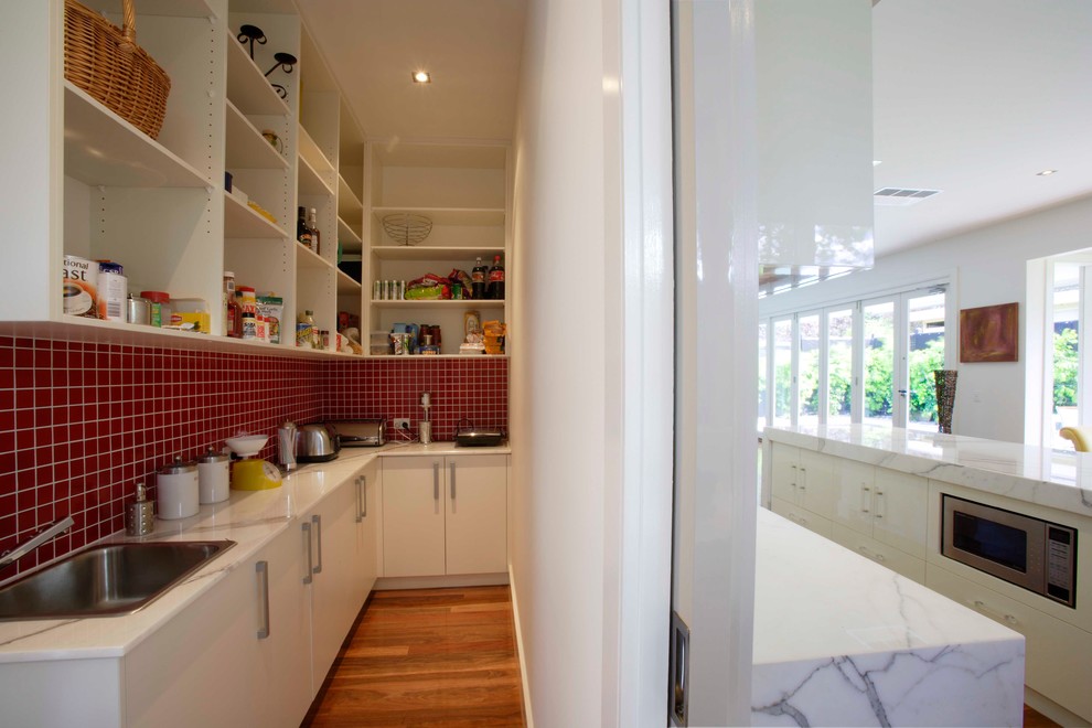 Kitchen pantry - large contemporary l-shaped medium tone wood floor kitchen pantry idea in Melbourne with an undermount sink, flat-panel cabinets, white cabinets, marble countertops, white backsplash, glass sheet backsplash, stainless steel appliances and an island