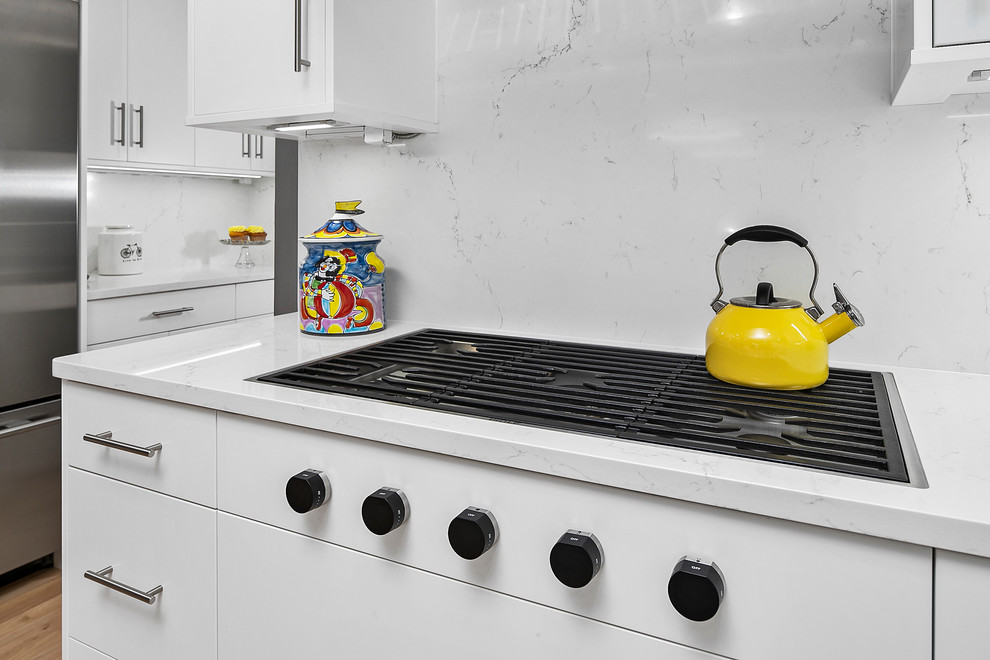 This is an example of a large contemporary l-shaped kitchen/diner in Detroit with a submerged sink, flat-panel cabinets, white cabinets, engineered stone countertops, white splashback, stone slab splashback, stainless steel appliances, medium hardwood flooring, an island, brown floors and white worktops.