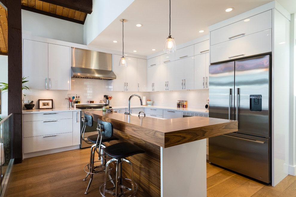 Medium sized contemporary l-shaped kitchen in Vancouver with flat-panel cabinets, white cabinets, engineered stone countertops, white splashback, stainless steel appliances, an island, brown floors, metro tiled splashback and medium hardwood flooring.